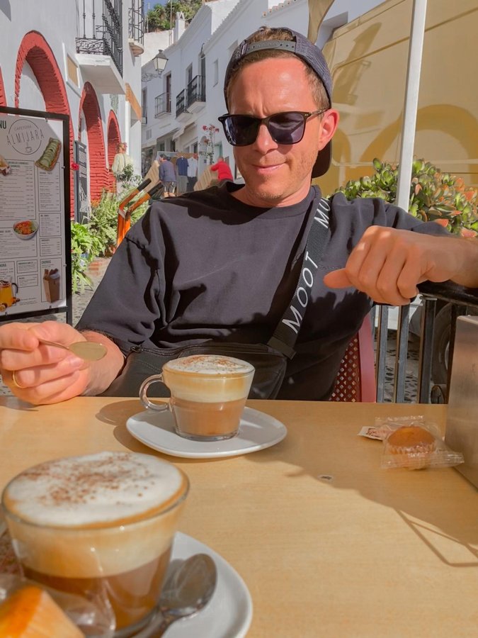 André mit Sonnenbrille an einem sonnigen Cafétisch, vor ihm zwei Cappuccini – er zeigt auf eine der Tassen